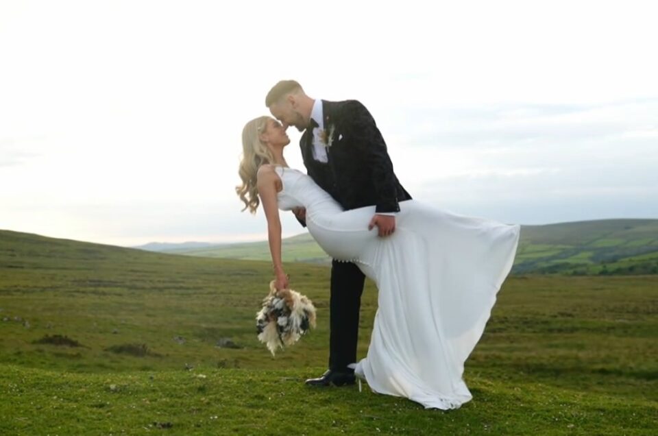 Groom in black tie dripping blonde bride in a welsh landscape. Bride is in a flowing dress holding a buoqet,