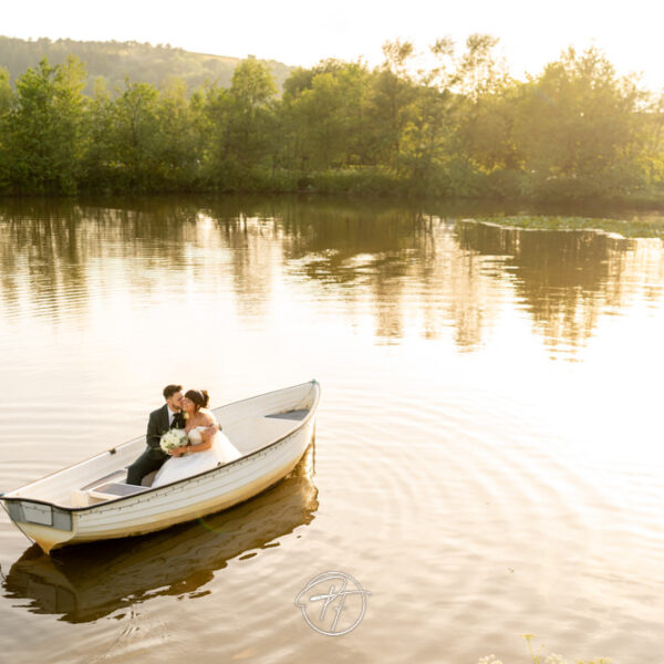 Wide-angle drone or high-angle view of a bride and groom sitting in a white rowing boat on a calm lake during golden hour at Sylen Lakes in Llanelli. Photography First.