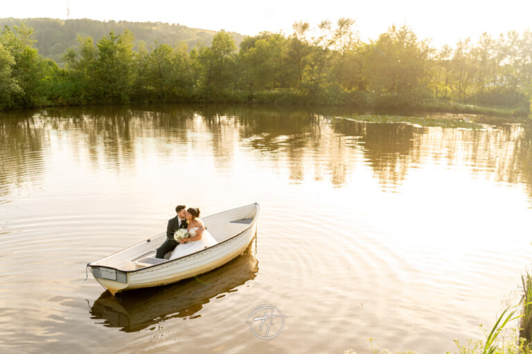 Wide-angle drone or high-angle view of a bride and groom sitting in a white rowing boat on a calm lake during golden hour at Sylen Lakes in Llanelli. Photography First.