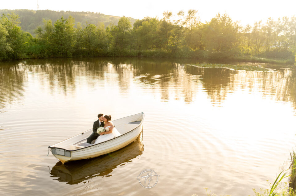 Wide-angle drone or high-angle view of a bride and groom sitting in a white rowing boat on a calm lake during golden hour at Sylen Lakes in Llanelli. Photography First.